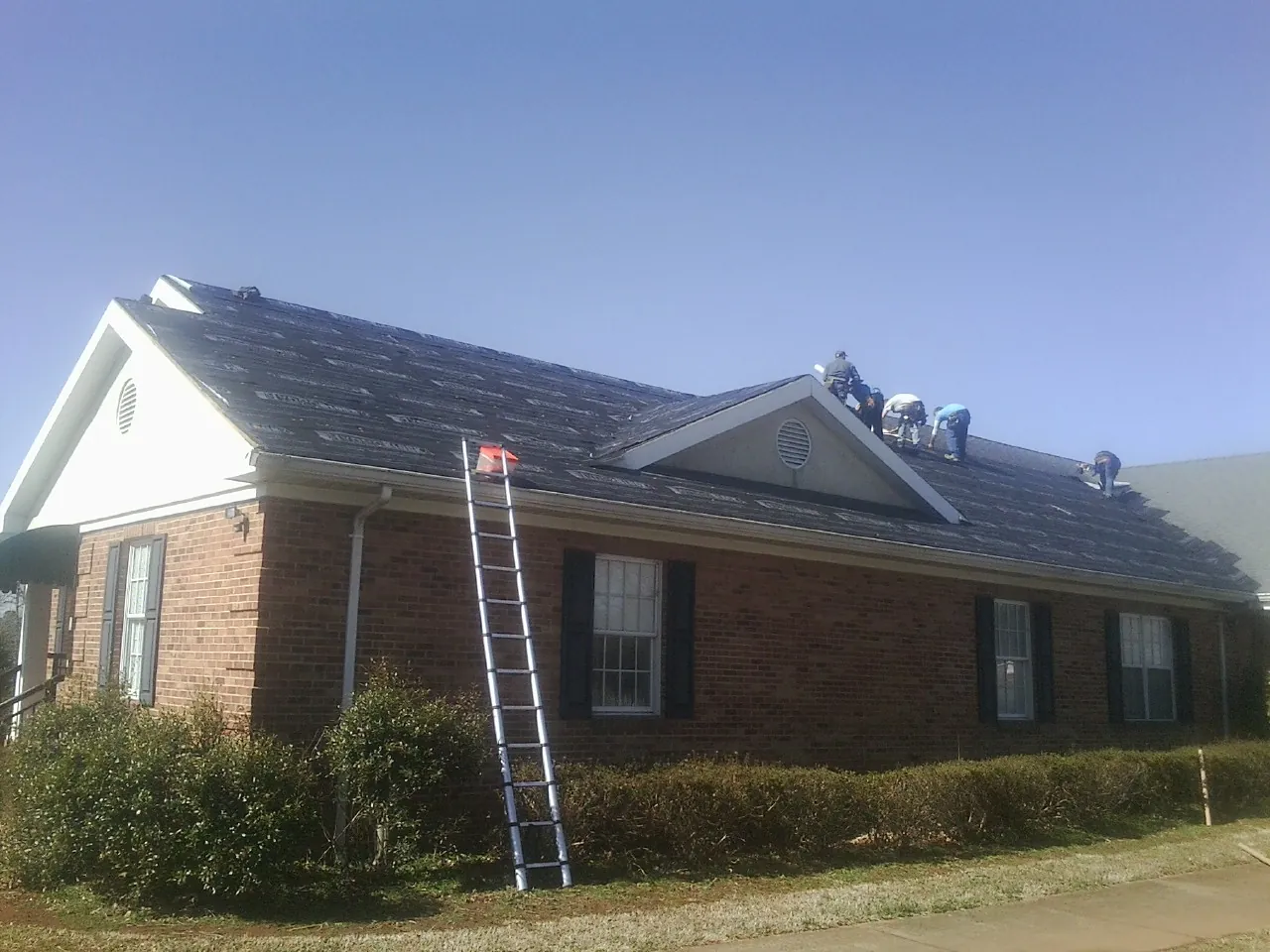 Crew tearing shingles off a brick ranch roof with a ladder set — Metal Roof Repair work by our crew in Nunda